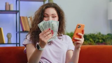 Aile bütçesi planlamak. Gülümseyen genç bir kadın nakit para sayıyor, akıllı telefon kullanıyor ev faturalarını hesaplıyor. Neşeli kız gelirinden memnun ve planlı tatil ve hediyeler için para biriktiriyor.