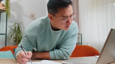 Hüzünlü Asyalı serbest çalışan evde deftere not alıyor, dizüstü bilgisayar kullanıyor, iş raporlarını hazırlıyor. Çevrimiçi hataları analiz eden muhasebeci vergileri, iflası, borcu hesaplar.