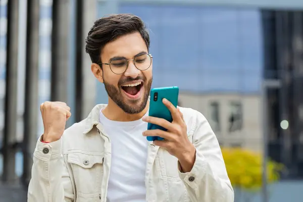 Joyful Indian man use mobile smartphone celebrating win good message news lottery jackpot victory, giveaway online outdoors. Happy Arabian Hindu guy in downtown city street. Business people lifestyles