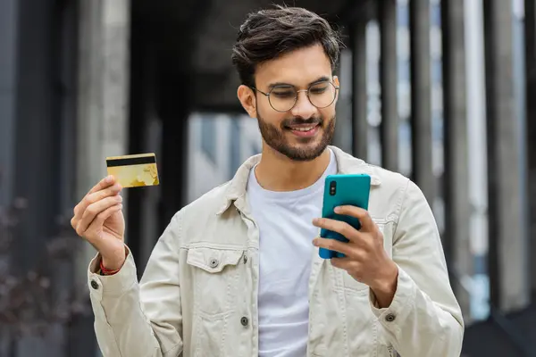 Joyful Indian man using credit bank card smartphone while transferring money, purchases online shopping order food delivery booking outdoors. Arabian Hindu guy in downtown city street. Business people