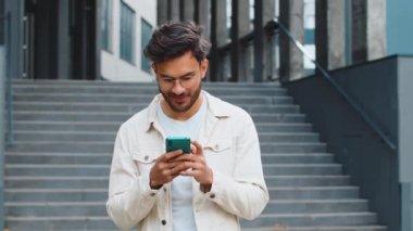 Genç gülümseyen Hintli adam akıllı telefon kullanıyor kısa mesajlar yazıyor, e-posta okuyor, dışarıda haberleri inceliyor. Şehir merkezinde yürüyen Arap Hindu adam mobil sosyal medya uygulaması kullanıyor. İş adamları