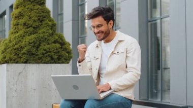 Dizüstü bilgisayarla çalışan mutlu Hintli genç adam sevinçle çığlık atıyor zafer jesti olarak ellerini kaldırıyor başarıyı kutluyor açık havada para kazanıyor. Arap Hintli adam şehir merkezindeki bankta oturuyor.