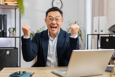 Dizüstü bilgisayarda çalışan mutlu, şaşkın, orta yaşlı Asyalı işadamı ani zafer, maç kazanma, piyango hedefi başarısı kariyeri iyi haberlerle şok oldu. Ev ofis çalışma masasında heyecanlı bir serbest çalışan.