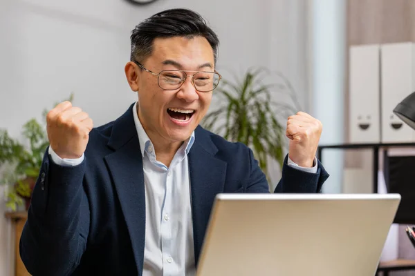 Happy Asian middle-aged business man working on laptop at home office desk typing browsing celebrating success victory, winning lottery jackpot goal achievement play game good positive news, triumph