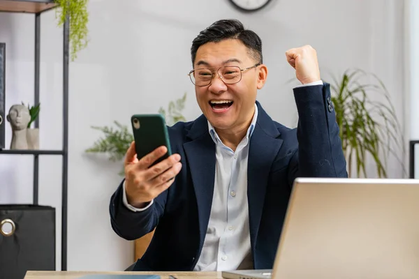 Happy Asian mature businessman freelancer use mobile smartphone celebrating success victory, winning lottery jackpot achievement, play game, good triumph positive news working at home office workplace