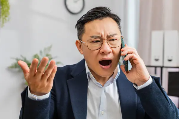 Conflict Quarrel Negotiation. Angry businessman CEO making smartphone online conversation call at office. Upset mad Asian man talking colleague after analyzing financial report mistake problem