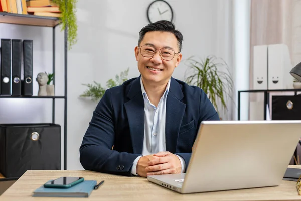Portrait of happy smiling Asian businessman at modern home office workplace desk looking at camera. Male Chinese in formal suit. Handsome middle-aged man remote distant online work on laptop notebook