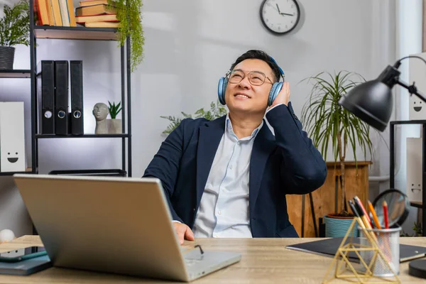 Happy relaxed overjoyed Asian businessman working on laptop computer at home office wearing headphones listening favorite energetic disco music and dancing. Freelancer man relaxing, taking a break