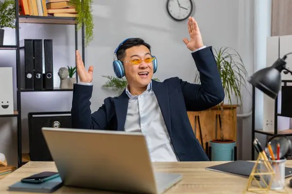 Happy relaxed Asian businessman in sunglasses working on laptop computer at home office wearing headphones listening favorite energetic disco music and dancing. Chinese man relaxing, taking a break