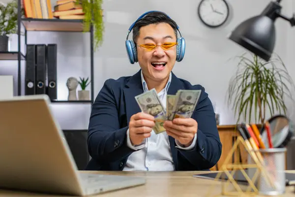 Asian businessman holding fan of cash money dollar banknotes celebrate, dance, success career, lottery jackpot game winner, big income, wealth at home office workplace desk. Remote distant working