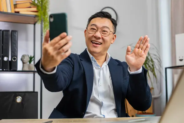 Happy excited Asian middle-aged businessman blogger influencer making phone conversation call with social media followers at office workplace working on laptop. Freelancer Chinese man in formal suit