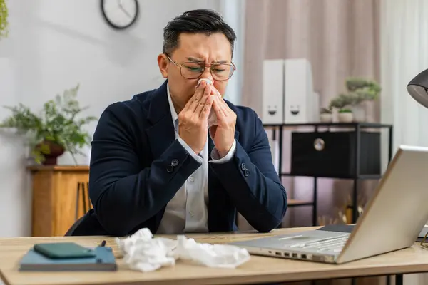 Sick ill Asian middle-aged businessman in suit suffers from cold flu fever or allergy working on laptop at home office. Overworked manager freelancer employee man blows sneezes wipes snot into napkin