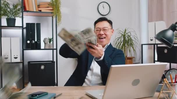 ハッピーリッチなアジアの中年ビジネスマンは 大きな利益を勝ち取る宝くじジャックポット成功した支払い購入キャッシュバックをオフィスでラップトップで働く周りにドル現金を共有することを投げます 中国人男性 — ストック動画
