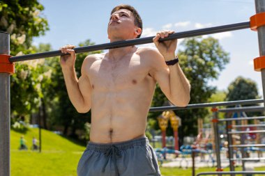 Beyaz atletik, üstsüz, kaslı adam yatay çubukta barfiks egzersizi yapıyor. Yazın oyun bahçesinde kaslarını şişiren adam. Spor sağlığı rutin egzersizi. Vücut geliştirme motivasyonu