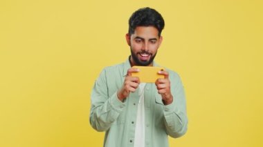 Endişeli Hintli sakallı adam heyecanlı bir şekilde yarış simülatörü ya da akıllı telefondan video oyunu oynuyor. Mutlu Arap Hindu oyuncu sarı stüdyo arka planında izole edilmiş zaferi kutluyor