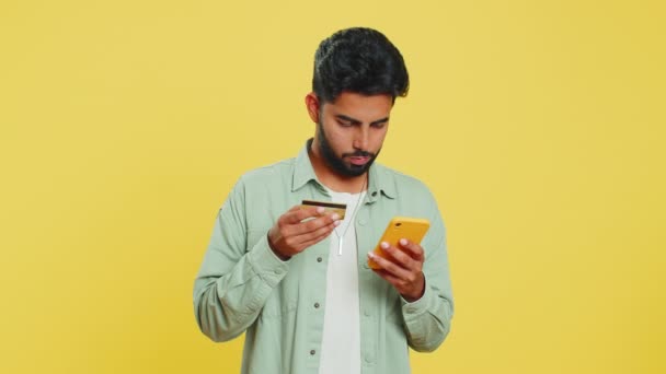 快乐的印度年轻人一边用信用卡和智能手机转移开销 一边在网上购物 订购食物 在黄色工作室背景上被隔离的阿拉伯人 — 图库视频影像