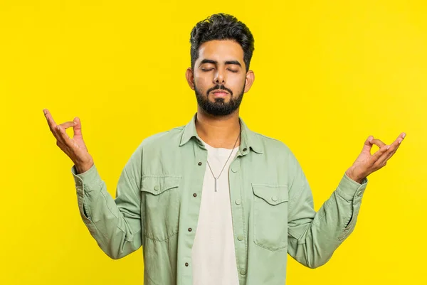 Keep calm down, relax, rest. Concentrated happy Indian man meditating breathes deeply with mudra yoga gesture, eyes closed, peaceful mind, taking a break. Arabian Hindu guy on yellow studio background