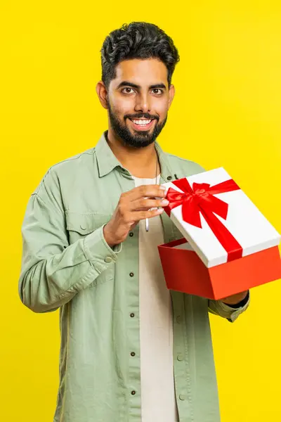 Happy Indian man opening birthday gift box with red ribbon. Holidays surprise concept. Smiling Arabian bearded guy receive great wrapped present celebrating isolated on yellow background. Vertical