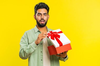 Mutlu Hintli genç adam kırmızı kurdeleli doğum günü hediyesi kutusunu açıyor. Tatil sürprizi konsepti. Gülümseyen Arap sakallı adam sarı stüdyo arka planında izole edilmiş büyük bir kutlama hediyesi aldı.