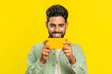 Endişeli Hintli sakallı adam heyecanlı bir şekilde yarış simülatörü ya da akıllı telefondan video oyunu oynuyor. Mutlu Arap Hindu oyuncu sarı stüdyo arka planında izole edilmiş zaferi kutluyor