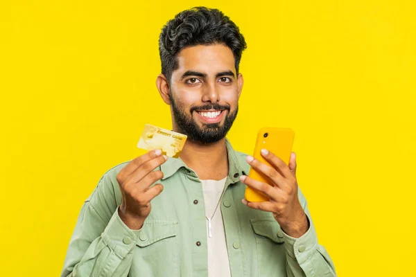 Happy young Indian man using credit bank card and smartphone while transferring spending money, purchases online shopping, ordering food delivery. Arabian guy isolated on yellow studio background