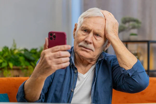 Senior sad man use smartphone typing browsing, loses becoming surprised sudden lottery results, bad news, fortune loss, fail. Elderly old grandfather at home living room sits on couch at table desk
