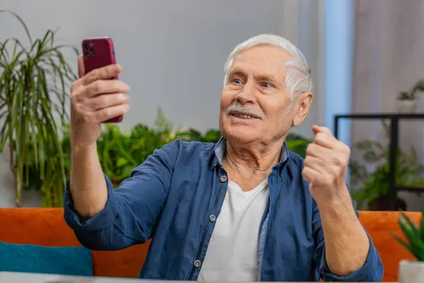 Happy senior man use mobile smartphone typing browsing celebrating success victory, winning lottery jackpot achievement play game good positive news at home. Elderly mature grandfather sits on couch