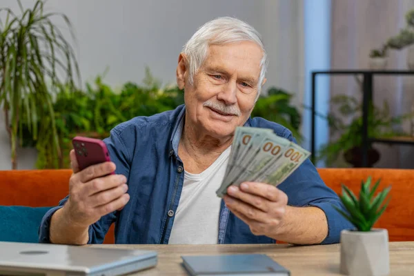 Planning family budget. Smiling senior man counting money cash use smartphone calculate domestic bills at home. Mature old grandfather satisfied of income and saves money for planned vacation gifts