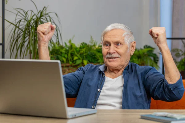 Excited senior old man at home table with laptop shouting in delight raise hands in triumph winner gesture celebrate success win money in lottery. Happy elderly mature grandfather get online good news