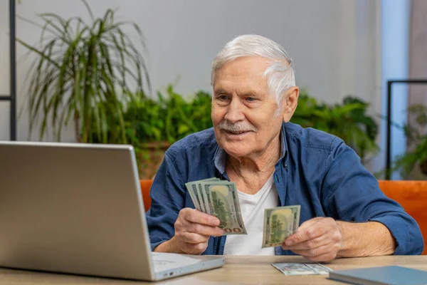 Rich happy senior old man counting money cash use laptop computer calculate domestic bills at home indoors. Elderly grandpa pensioner satisfied of income earnings, saves money for vacation and gifts