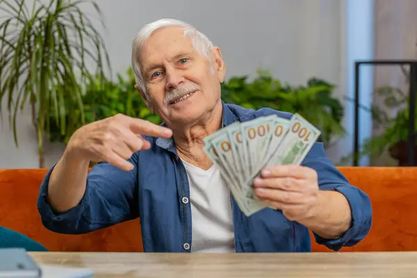 Planning family budget. Happy senior man counting money cash pointing finger on dollars at home. Elderly old grandfather satisfied of income salary pension and saves money for planned vacation gifts