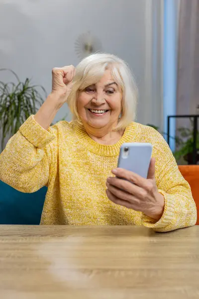 Happy senior old woman use mobile smartphone typing browsing celebrating success victory, winning lottery jackpot achievement play game good positive news at home. Elderly mature grandmother. Vertical