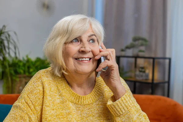 Phone call, good news, gossip. Happy excited senior old woman in pleasant conversation on smartphone, enjoying talking with friend at home apartment. Elderly mature grandmother sits on couch at table