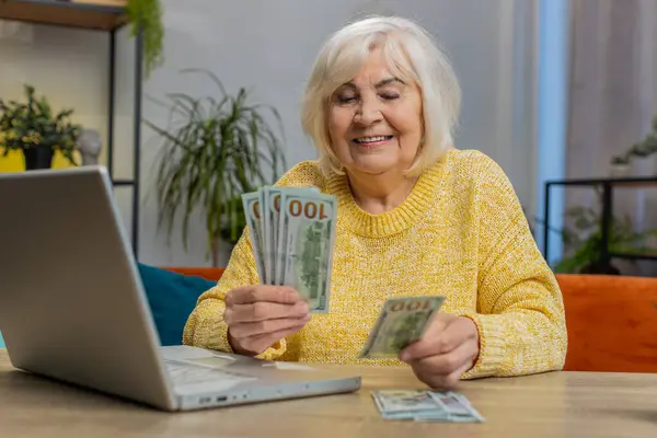 Rich happy senior old woman counting money cash use laptop computer calculate domestic bills at home room. Elderly grandmother pensioner satisfied of income earnings, saves money for vacation gifts