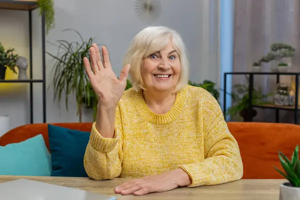 Hello. Senior old woman smiling friendly at camera waving hands gesturing hello, hi, greeting, goodbye, welcoming with hospitable expression at home indoors. Elderly mature grandmother sits at table