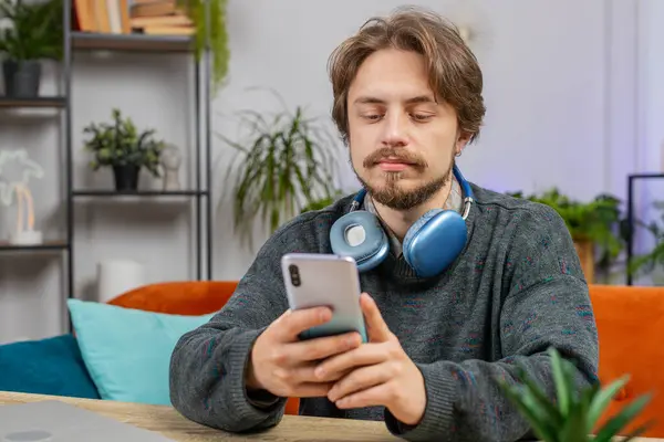 Caucasian man sits at table uses mobile phone smiles at home office room workplace. Young bearded guy texting share messages content on smartphone social media applications online watching relax movie