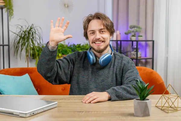 Hello. Bearded young man smiling friendly at camera and waving hands gesturing hello hi, greeting or goodbye welcoming with hospitable expression at home office. Caucasian guy sitting at table in room