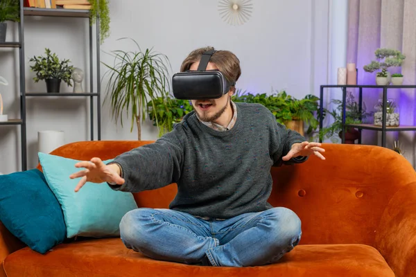Bearded man using virtual reality futuristic technology VR app headset helmet to play simulation 3D 360 online video game, watching film movie at modern home apartment. Guy in goggles sitting on couch