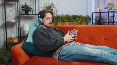 Kablosuz kulaklıklı mutlu, gevşemiş Kafkasyalı genç adam evinde kanepede dans ediyor ve akıllı telefon eğlencesinde favori disko müziğini dinliyor. İnsanlar hafta sonu aktiviteleri