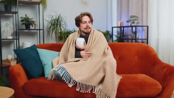 病気の男は 冷たい飲み物のホットコーヒーやハーブティーを熱せずに自宅のアパートで暖房しないで震えています 不健康なコーカサス人の男はソファーに座って暖かくしようとします — ストック動画