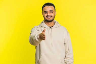 Kameraya uzanan dost canlısı Hintli genç adam, el sıkışma teklif ediyor, nazik bir gülümseme, davetiye, dayanışma ve güven göstergesi olarak karşılıyor. Sarı arka planda izole edilmiş Arap adam.