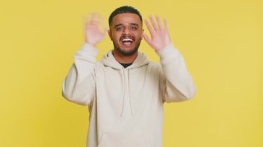 Kameraya dostça gülümseyen genç Hintli adam el sallayarak merhaba diyor ya da davetkar bir yüz ifadesiyle veda ediyor. Sarı stüdyo arka planında izole edilmiş Arap Hindu adam.