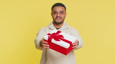 Pozitif gülümseyen Hintli adam doğum günü hediyelerini sunar ellerini uzatır, hediye paketi hediye ikramiyesi teklif eder, kutlama partisi düzenler, indirim indirimi yapar. Sarı arka planda izole edilmiş Arap adam.