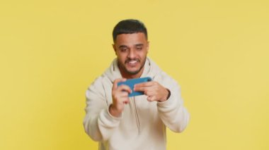 Endişeli Hintli sakallı adam heyecanlı bir şekilde yarış simülatörü ya da akıllı telefondan video oyunu oynuyor. Mutlu Arap Hindu oyuncu sarı stüdyo arka planında izole edilmiş zaferi kutluyor