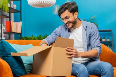 Paketleri boşaltan mutlu Hint Arap adam evde oturuyor. Mutlu gülümseyen Arap Hintli adam online alışveriş yapan dükkan müşterisi karton kutu açan müşteri hızlı posta yoluyla satın alma hediyesi alıyor