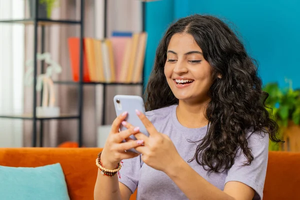 Indian woman sits on couch uses mobile phone smiles at home living room apartment. Arabian Hindu girl texting share messages content on smartphone social media applications online watching relax movie