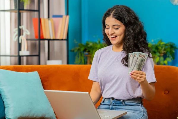 Planning budget. Rich happy Indian woman counting money cash use laptop computer calculate domestic bills at home. Arabian girl satisfied of income earnings, saves money for planned vacation, gifts