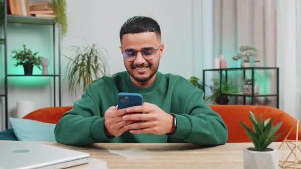 Hombre Indio Sienta Mesa Utiliza Sonrisas Teléfono Móvil Apartamento Oficina — Vídeos de Stock