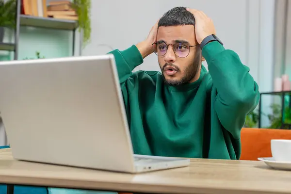 Displeased sad Indian man use laptop notebook typing browsing working, loses becoming surprised sudden lottery results bad news fortune loss game fail computer virus. Arabian guy at home office desk