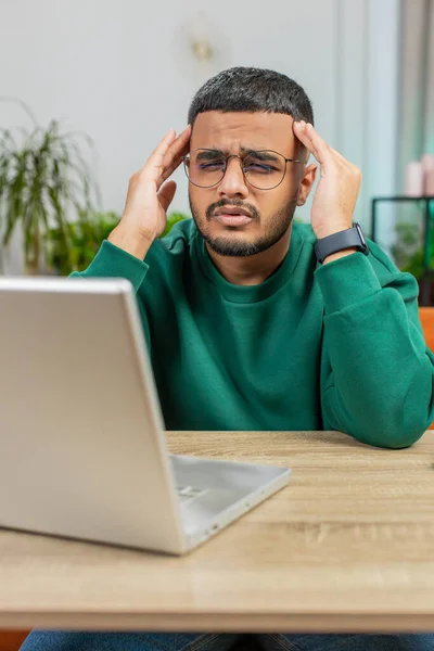 Tired Indian man freelancer use laptop, suffering from headache problem tension, migraine, stress at home office desk. Guy works on notebook, sends messages, makes online purchases, watching movies